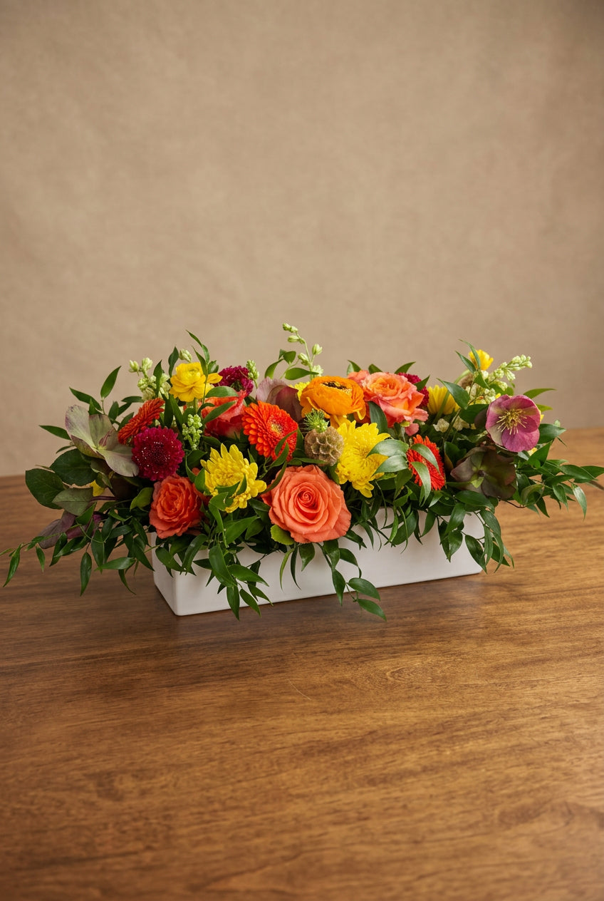 A vibrant, long rectangular floral arrangement featuring orange roses, yellow and red flowers, and green foliage, displayed in a white compote